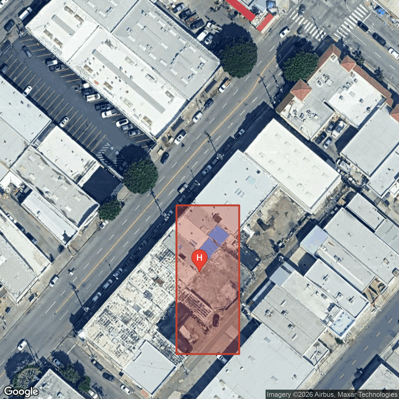Wider aerial centered on the corner of S San Pedro Street and Agatha Street with the approximate HCM-140 parcel boundary outlined in red and a placemarker on the building. The blue-tarp roof patch on the rear/east portion is clearly visible inside the polygon. Surrounding Flower District commercial buildings legible; the larger arterial visible at top is W 7th Street.