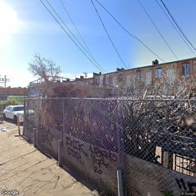 Alley / rear-elevation view of HCM-140 from behind a chain-link perimeter covered in graffiti. Visible: the rear brick walls with exposed and weathered sections, boarded windows, vegetation breaking through the chain-link, a worker observed on the roof attending to deteriorating sections, abandoned signage. This is the failing rear / east portion the audit's vision classifier flagged as 'distressed' and that motivated the ARCHITECTURALLY_SIGNIFICANT_AND_FAILING type-3 verdict.